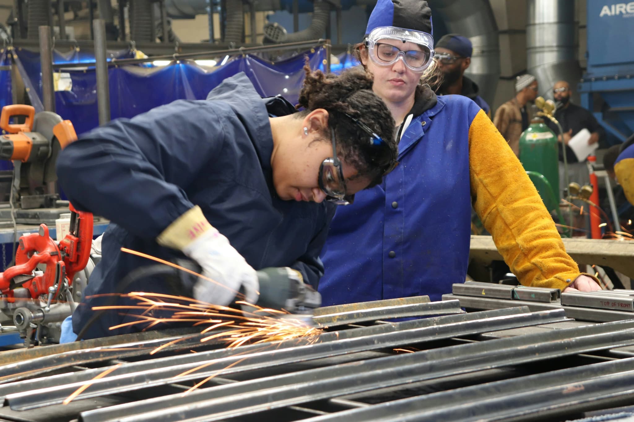 An apprentice cutting steel in a training shop while an instructor watches over their shoulder